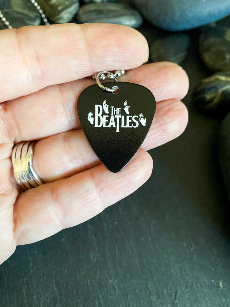 Beatles guitar pick necklace featuring their faces and the Beatles logo in white on a black guitar pick. Stainless steel ball chain. Band merch at Rock & Roll Jane. Held in a hand.