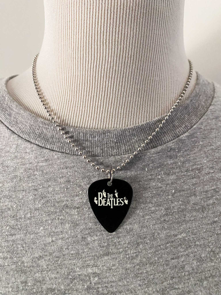 Beatles guitar pick necklace featuring their faces and the Beatles logo in white on a black guitar pick. Stainless steel ball chain. Band merch at Rock & Roll Jane. Displayed on a mannequin.