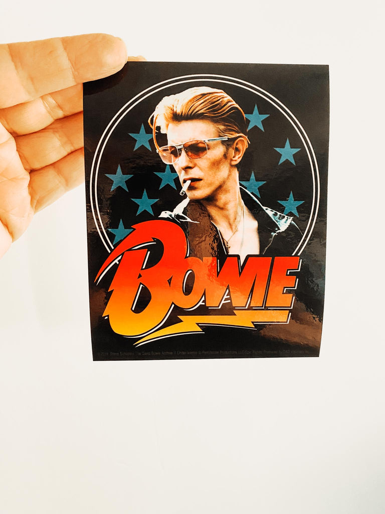 Officially licensed David Bowie stars sticker. Picture of David Bowie with sunglasses and a cigarette in his mouth. Blue stars in the background. The word "Bowie" in red.