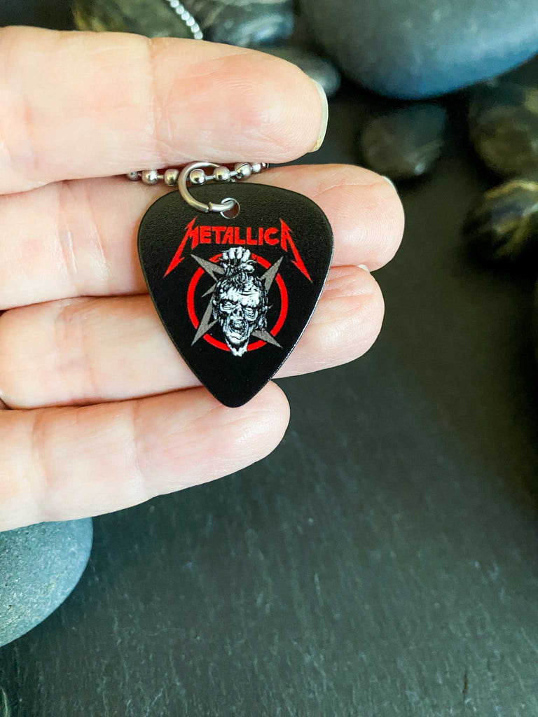 Metallica guitar pick necklace featuring the artwork from Damage, Inc. with a skull and the band's name in red. Band merch at Rock & Roll Jane. Held in someone's hand.