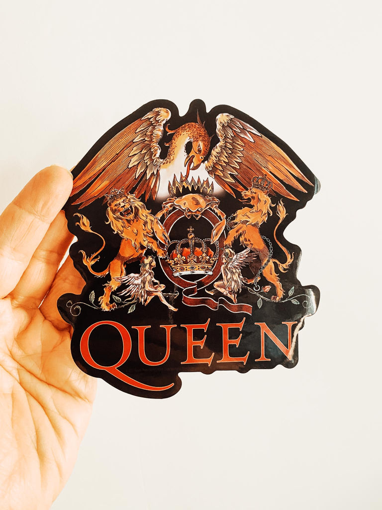 Sticker with 'QUEEN' and emblem design held by a hand on a white background. Officially licensed QUEEN classic crest sticker available at Rock & Roll Jane.