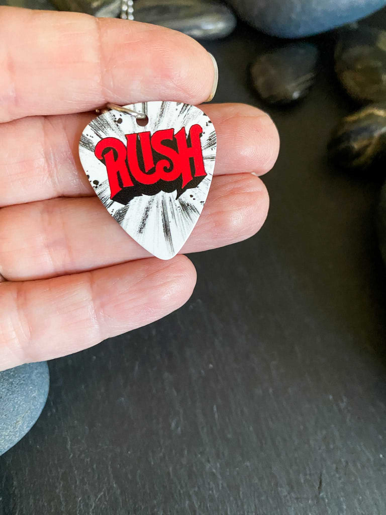 Rush guitar pick necklace on an 18" stainless steel ball chain. Band merch available at Rock & Roll Jane. Red logo lettering on a white and black background. Guitar pick is being held in a hand.